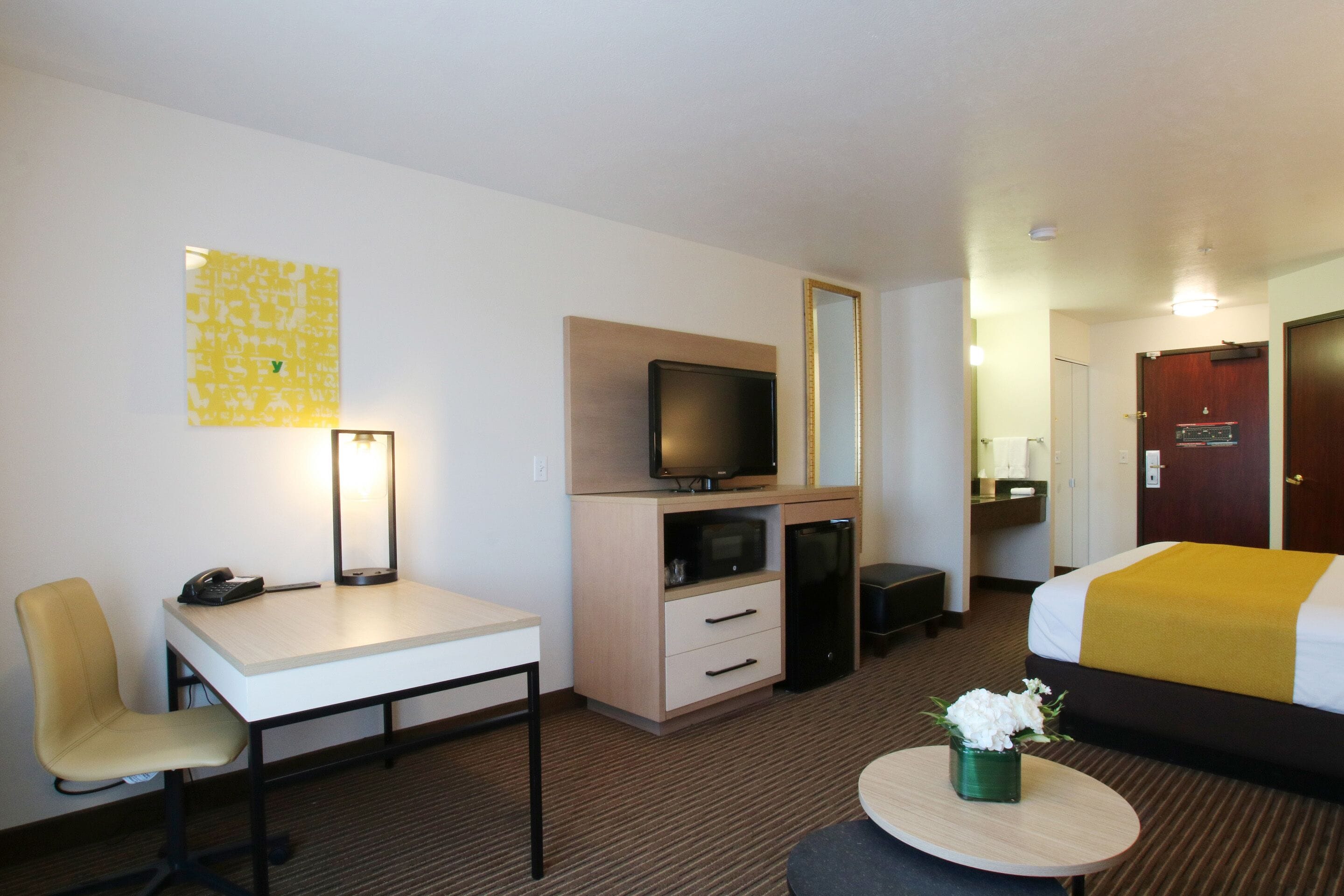 A hotel room with a bed, a desk with a chair, a television, a small refrigerator, and a bedside table with a vase of flowers.