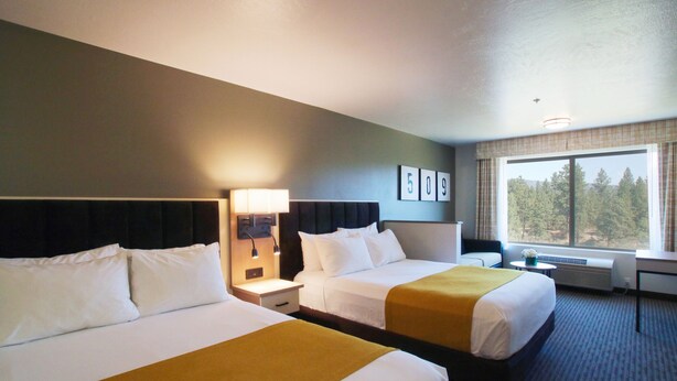 A hotel room with two beds, a nightstand, a lamp, a window with a view of trees, and a wall with a number sign. - Oxford Suites Spokane Valley (Spokane Valley)