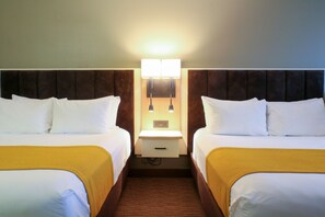 Quarto de hotel com duas camas, uma mesinha de cabeceira e um ponto de luz fixado na parede.