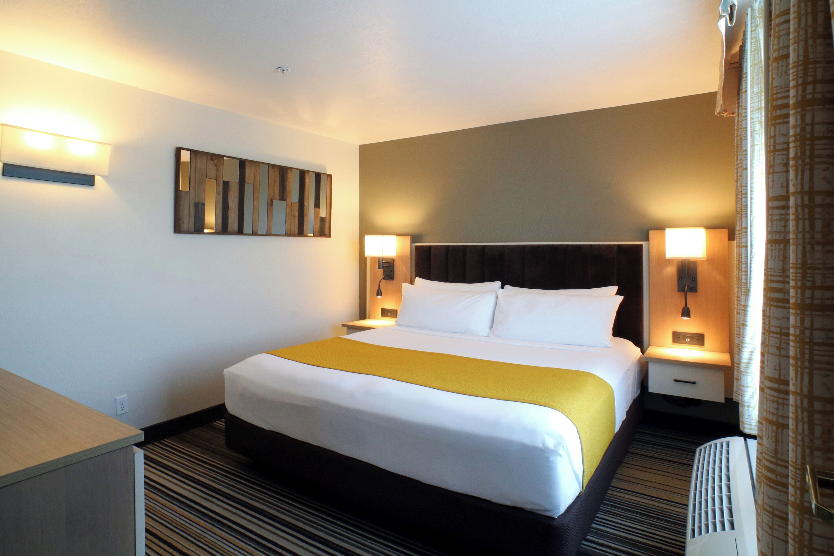 A hotel room with a large bed, two bedside tables, a nightstand, a wall-mounted mirror, and a window with curtains.