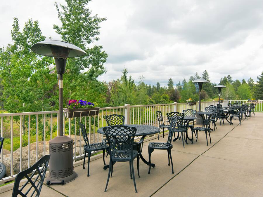 An outdoor patio with tables and chairs, surrounded by a railing, overlooking a landscaped area with trees and flowers.
