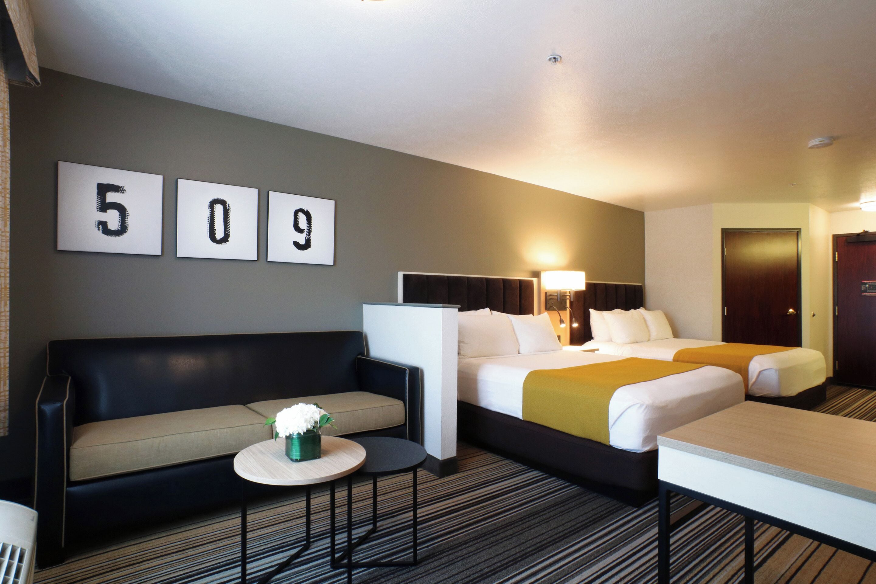 A hotel room with two beds, a sofa, a small table with a vase, and a wall with room numbers.