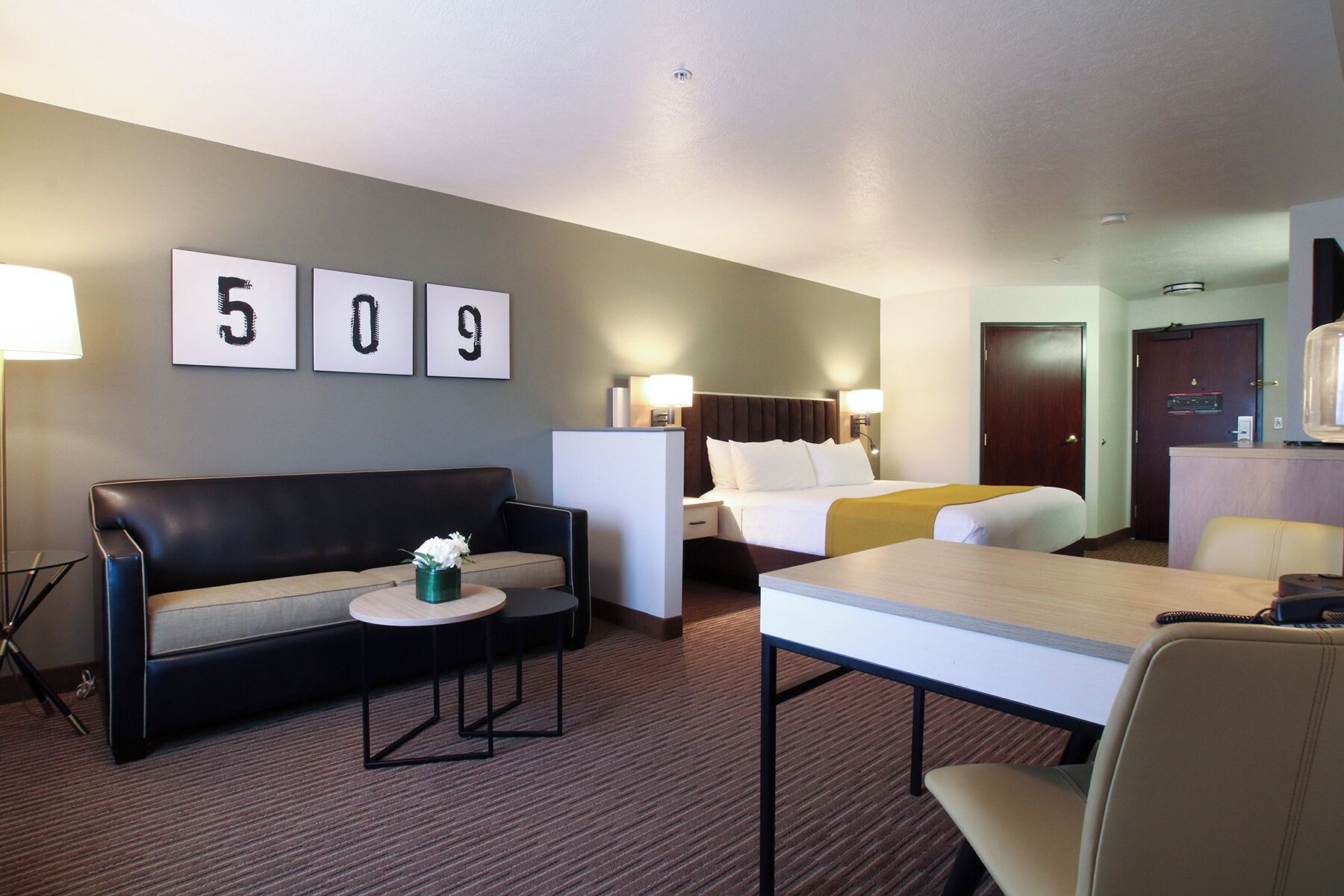 A hotel room with a bed, a sofa, a table, and a desk. There are wall-mounted lamps and a door.