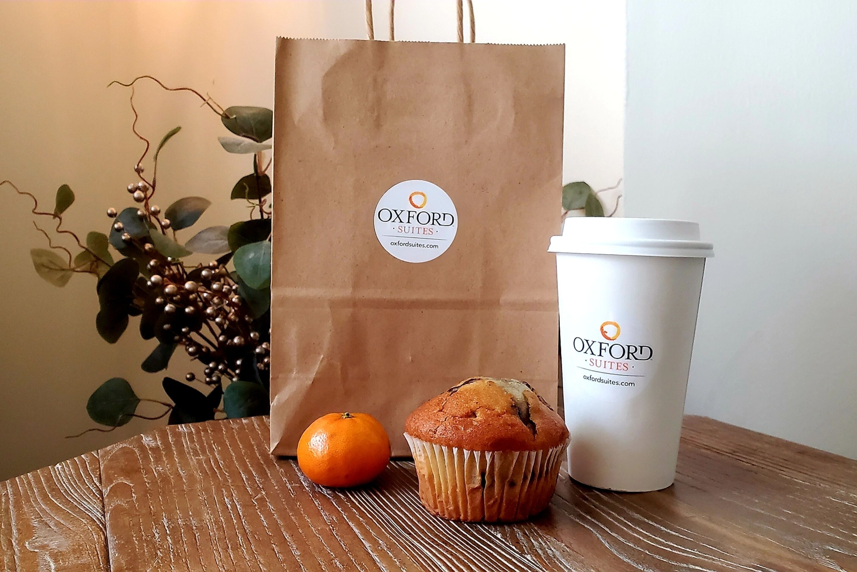 A brown paper bag and a white cup with the Oxford Suites logo, a muffin, and an orange on a wooden table.