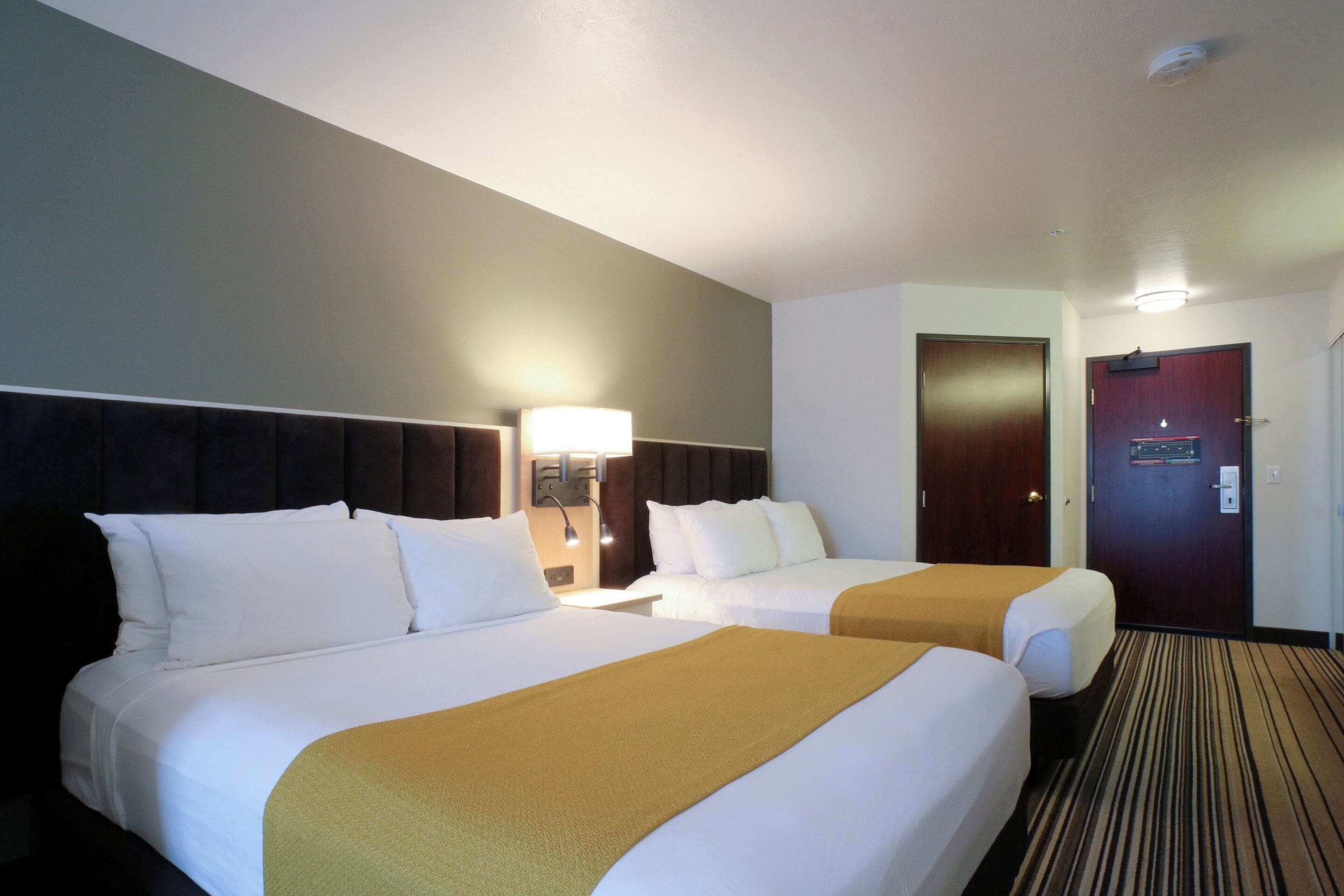 A hotel room with two beds, a nightstand with a lamp, a door, and a carpeted floor.
