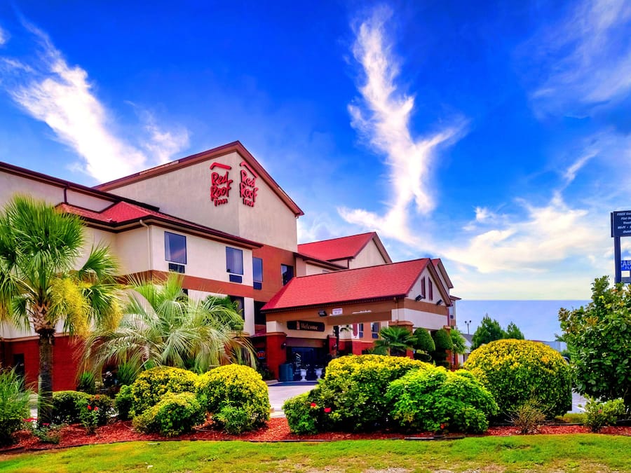 Un Red Roof Inn con techo rojo y paredes color beige, rodeado de jardines bien cuidados y palmeras.
