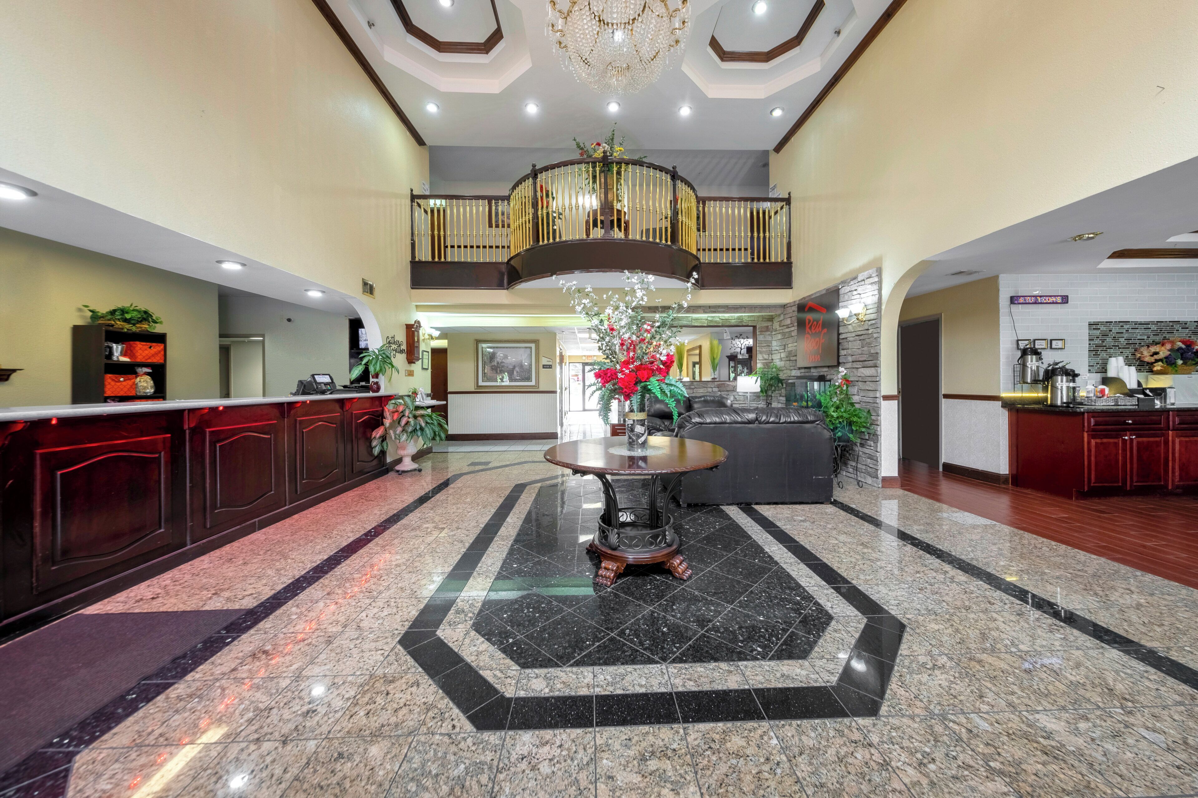 Een hotel lobby met een receptiebalie, een centrale tafel met een bloemenvaas, een kroonluchter en een trap naar een hoger niveau.