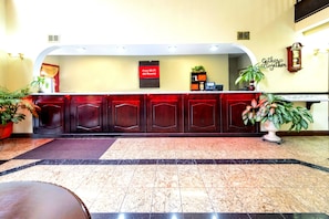 A reception area with a marble floor, wooden reception desk, and potted plants. - Red Roof Inn Atlanta Southeast - Stockbridge (Stockbridge)