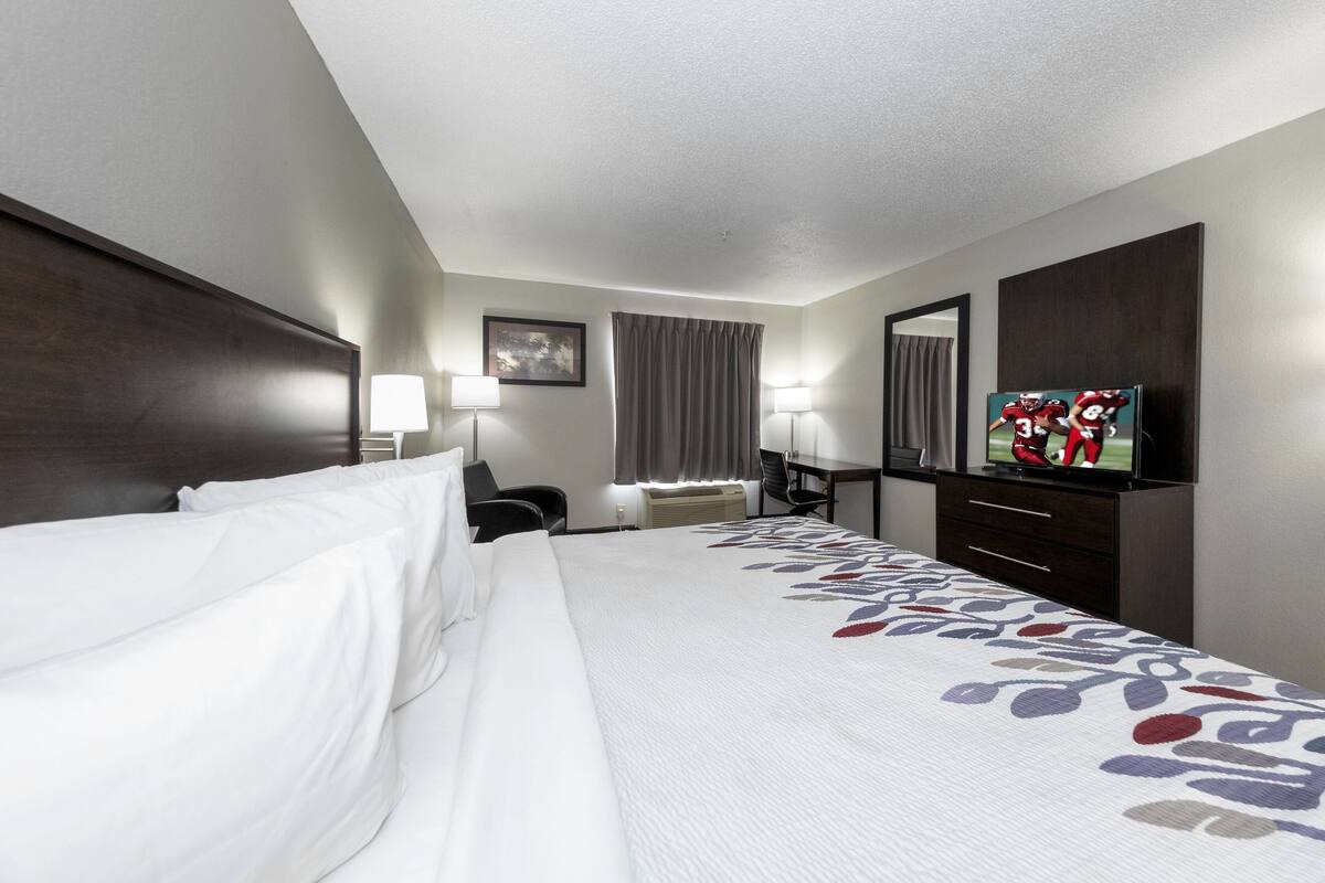 Une chambre d’hôtel avec un grand lit, un bureau avec une chaise, une télévision, une fenêtre avec des rideaux et un miroir fixé au mur.
