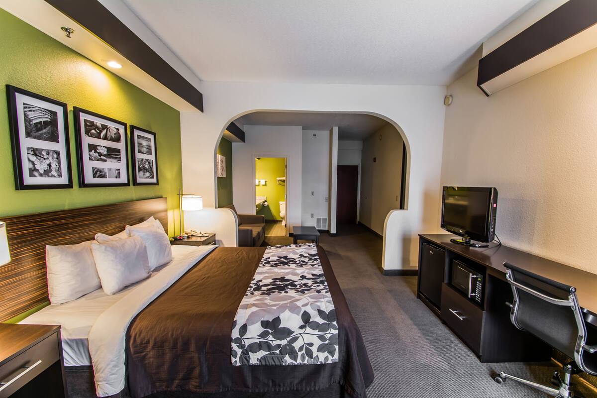 A hotel room with a bed, a desk, a TV, and a bathroom visible through an archway.