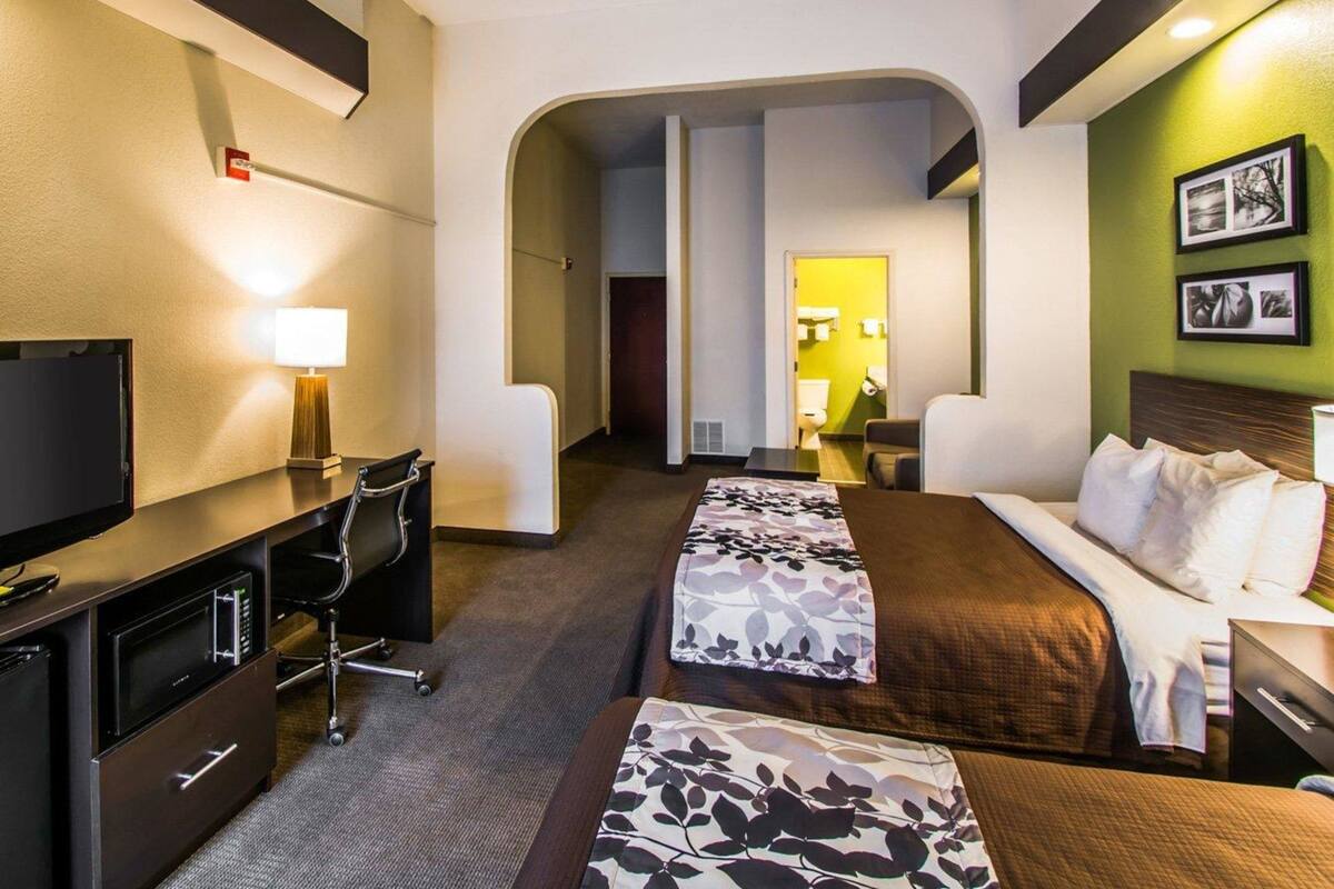 A hotel room with a bed, a desk, a chair, a television, and a bathroom visible through an archway.
