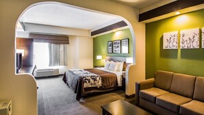 A hotel room with a bed, a sofa, a coffee table, and a desk. There is a window with curtains, a wall with framed pictures, and a green accent wall.