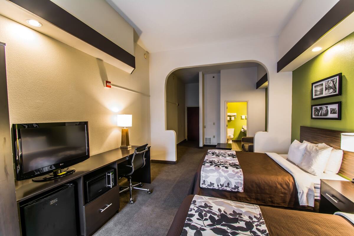 A hotel room with a flat-screen TV, a desk with a lamp, a microwave, a refrigerator, a bed with floral bedding, and a headboard with a lamp.
