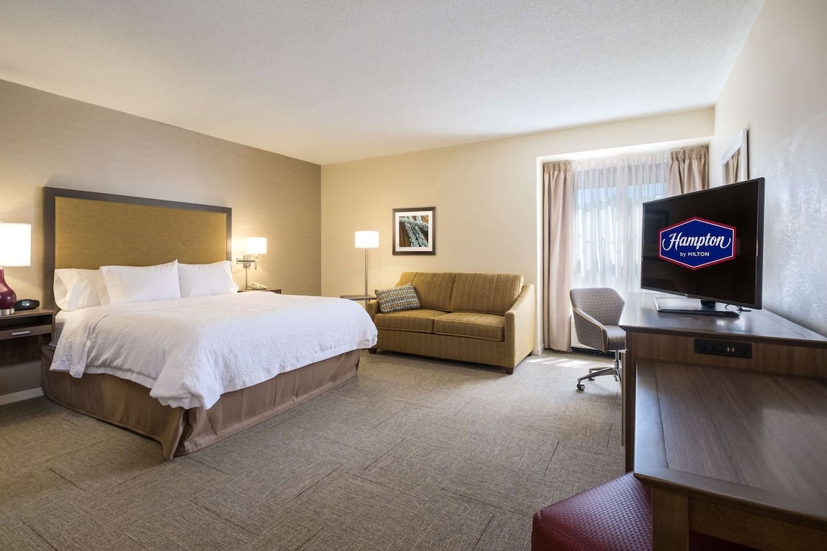 A hotel room with a bed, a sofa, a desk with a television, and a window with curtains.