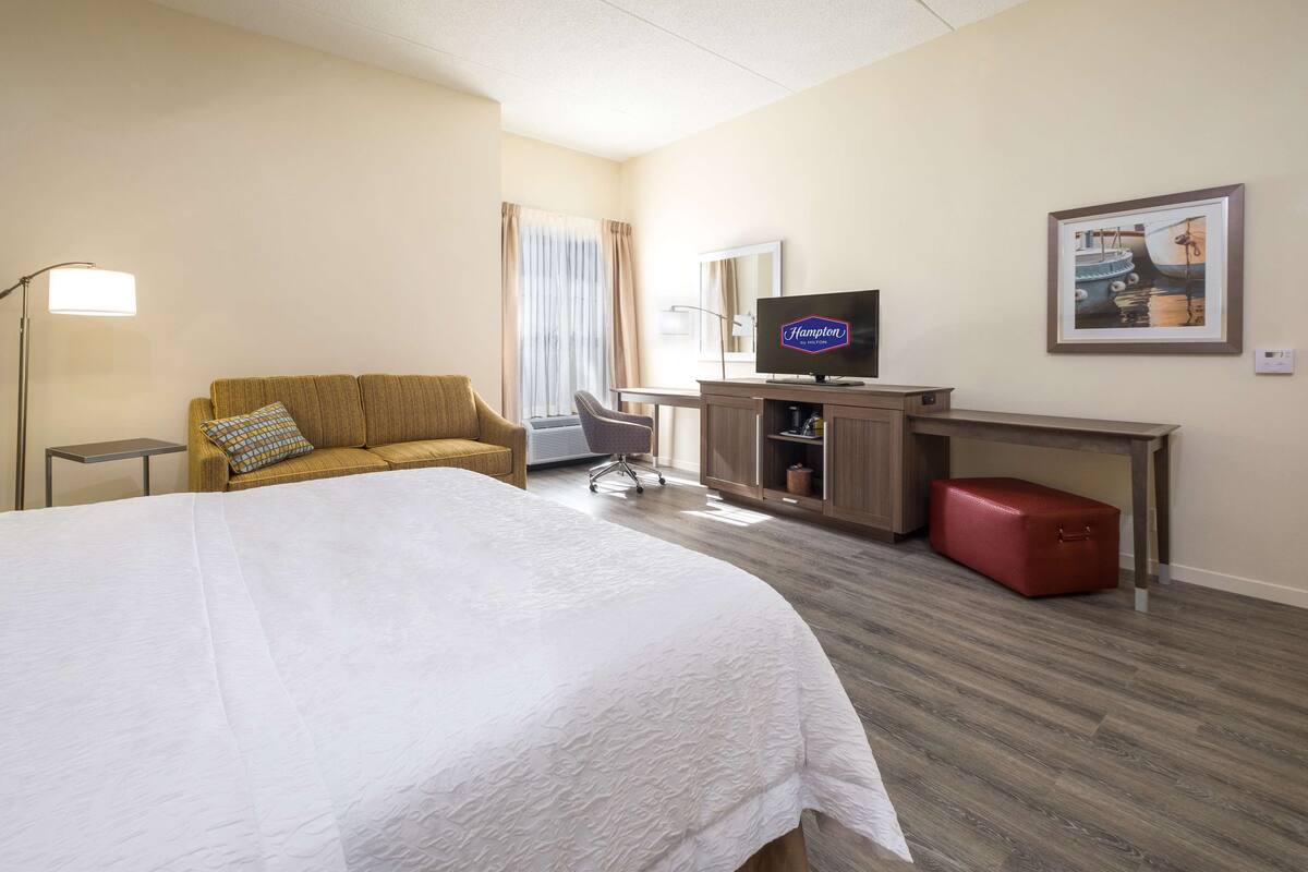 A hotel room with a large bed, a sofa, a desk with a television, a red suitcase, and a framed picture on the wall.