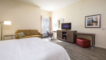 A hotel room with a large bed, a sofa, a desk with a television, a red suitcase, and a framed picture on the wall.