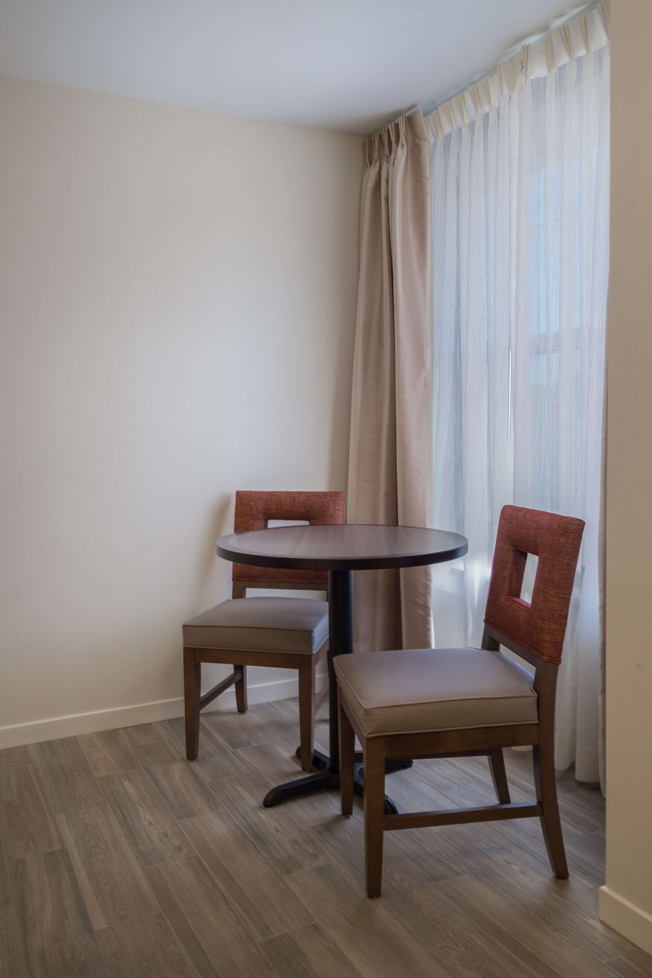 A corner room with a round table and two chairs, wooden flooring, and a window with sheer curtains.