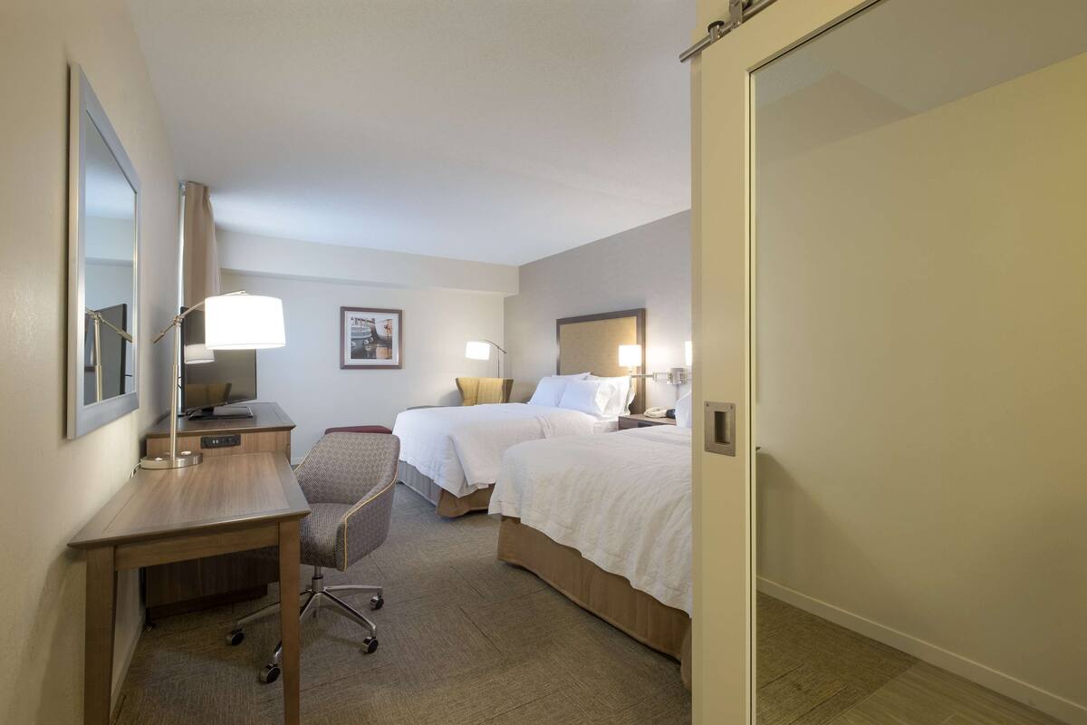 A hotel room with two beds, a desk, a chair, and a mirror.