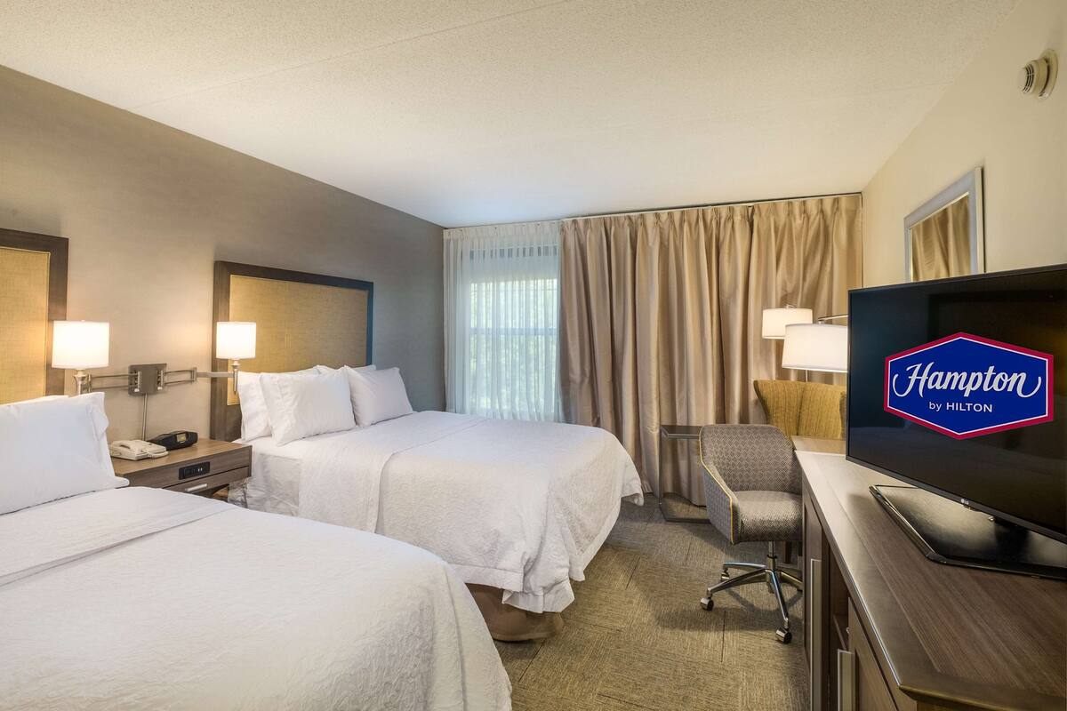 A hotel room with two beds, a desk with a television, a chair, and a window with curtains.