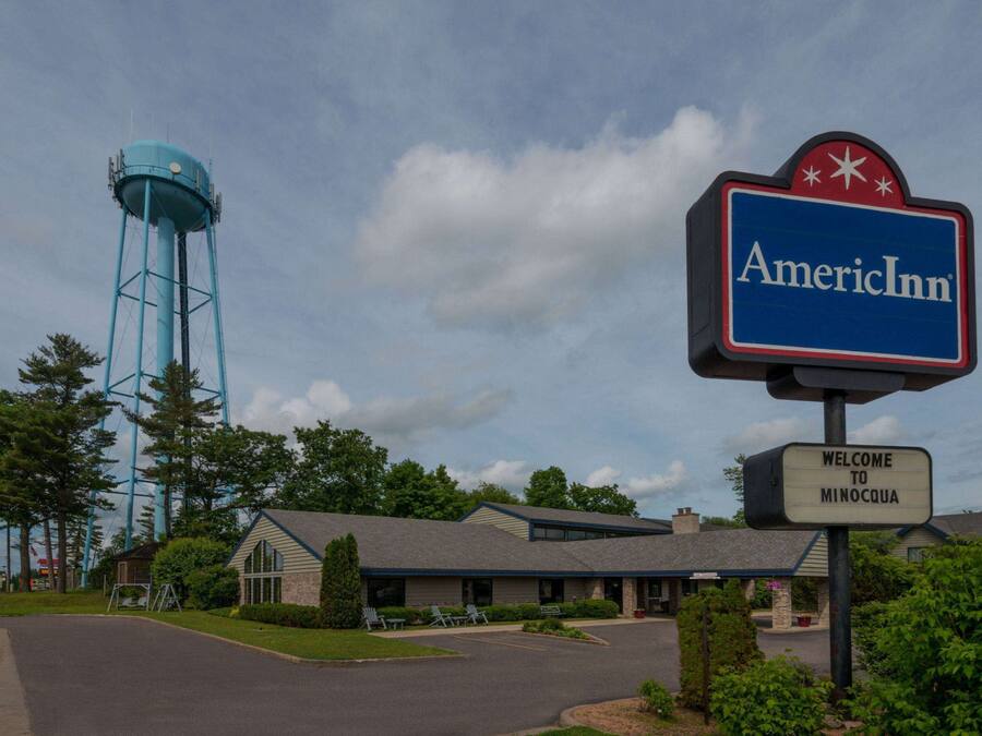 Letrero de hotel con el texto "AmericInn" y "Bienvenido a Minocqua", una torre de agua y un edificio con techo inclinado.