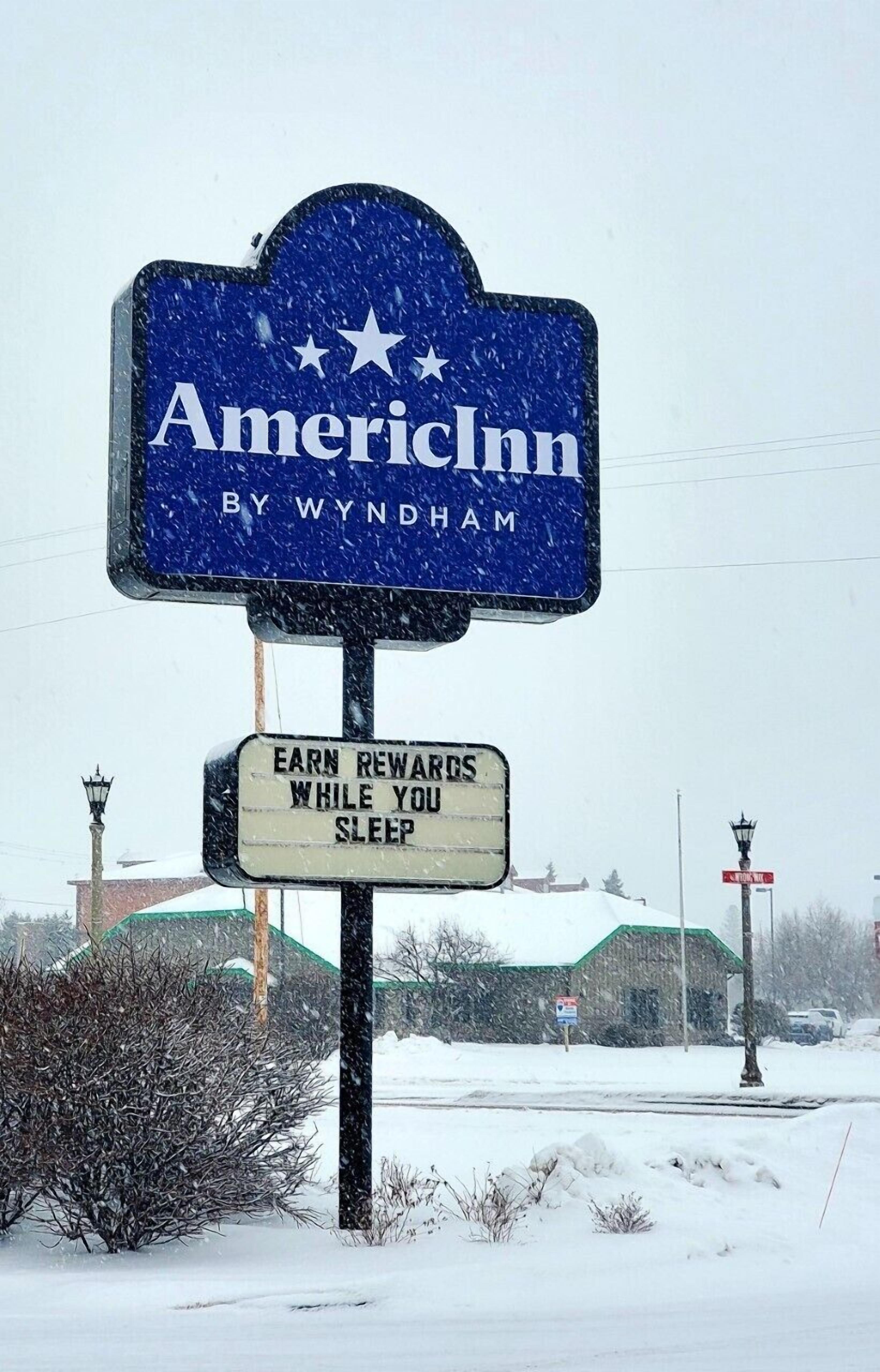 A snow-covered sign for AmericInn by Wyndham with a smaller sign below stating "Earn rewards while you sleep."