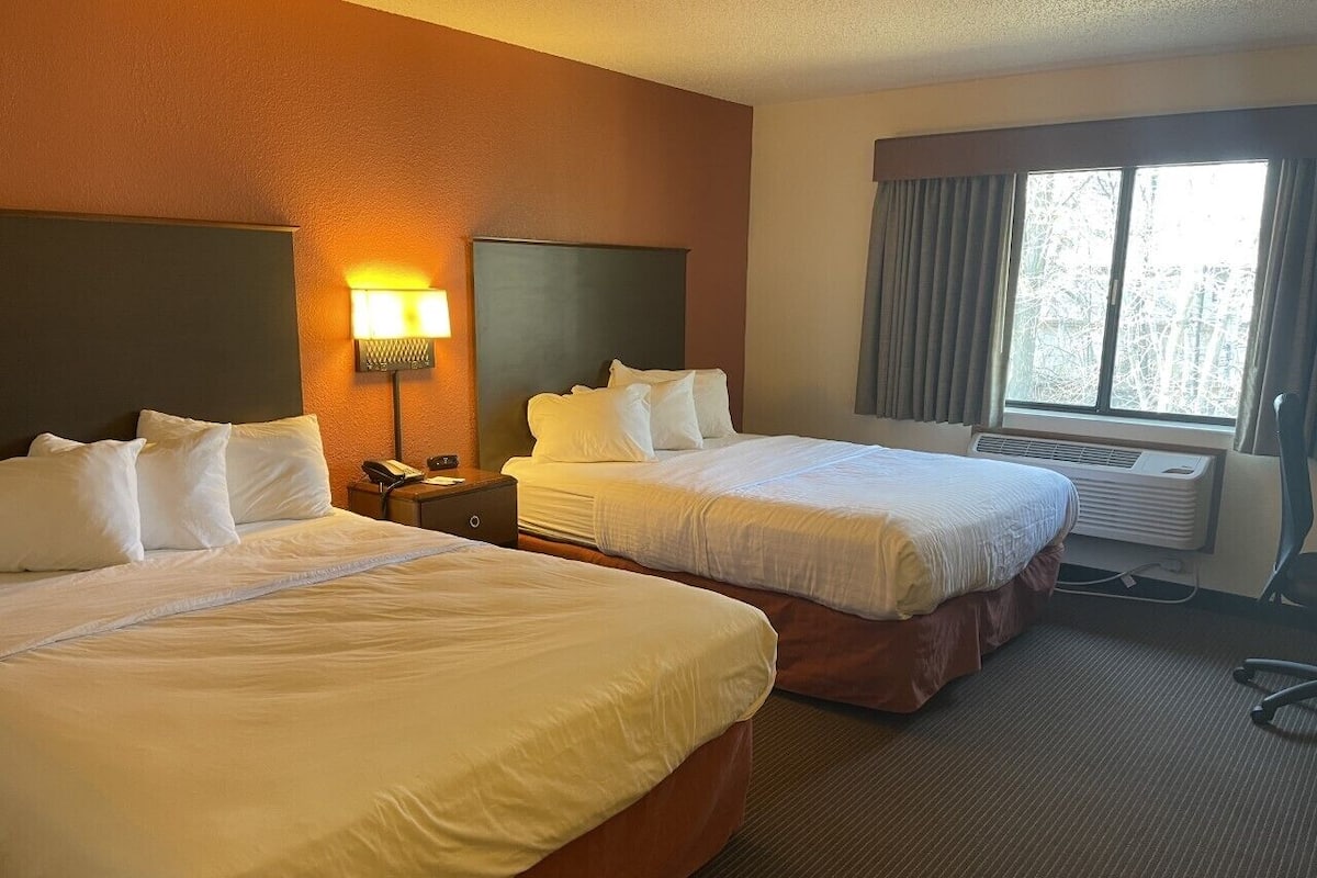Habitación de hotel con dos camas, una mesita de noche, una lámpara, una ventana con cortinas y un equipo de aire acondicionado.