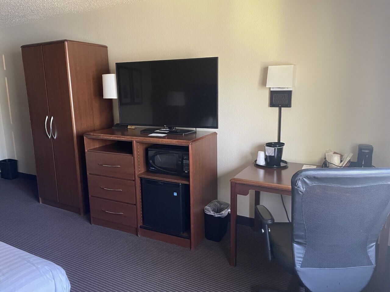 Habitación de hotel con cama, armario de madera, televisor, escritorio con lámpara, microondas, refrigerador y cesto de basura.