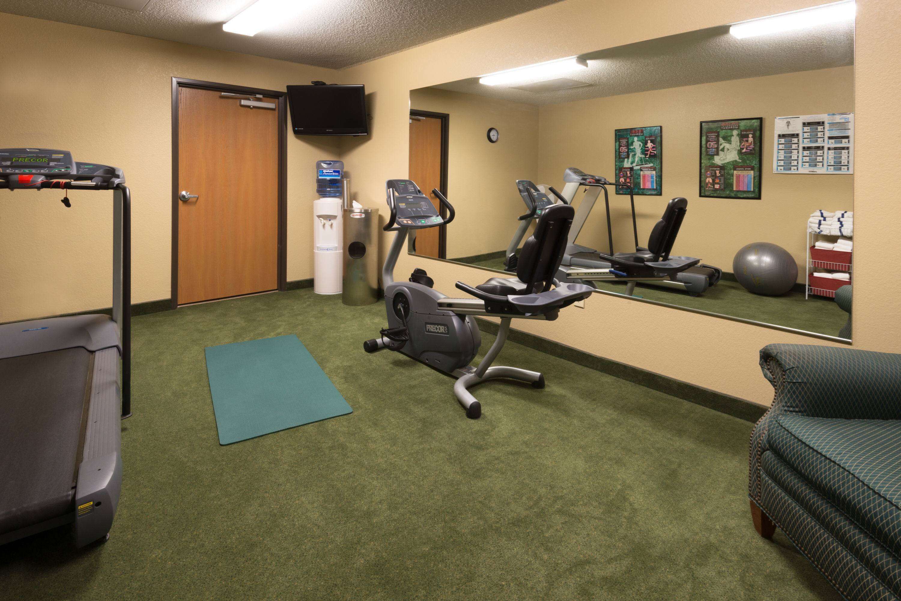Een sportschool met trainingsapparatuur, een loopband, een crosstrainer, een hometrainer en een yogamat.