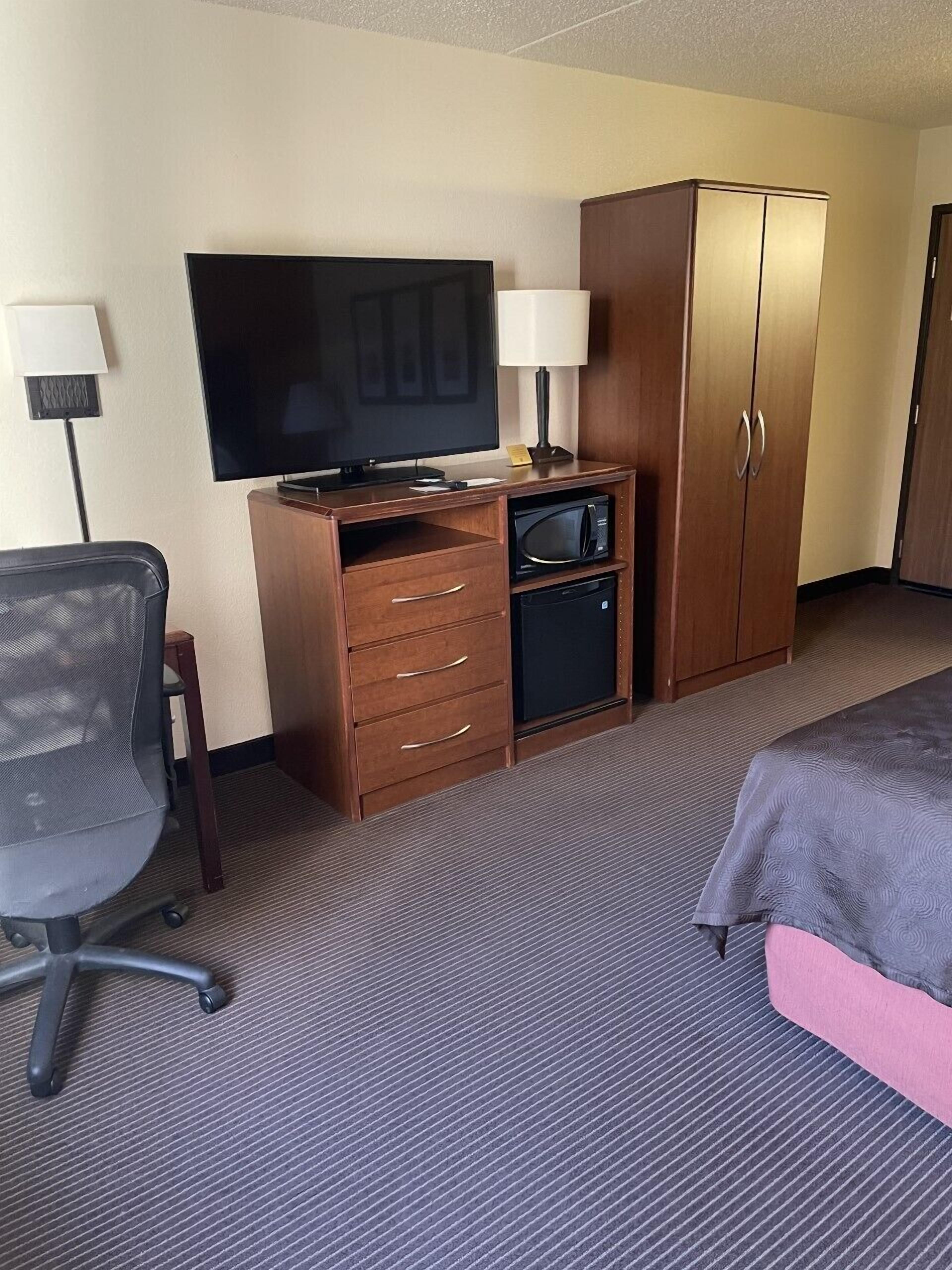 Ein Hotelzimmer mit Flachbildfernseher, Schreibtisch mit Lampe, Kleiderschrank, Mikrowelle, kleinem Kühlschrank, Bürostuhl und einem Bett.