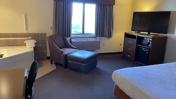 A hotel room with a bed, a chair with an ottoman, a desk, a television, a microwave, and a window with curtains.