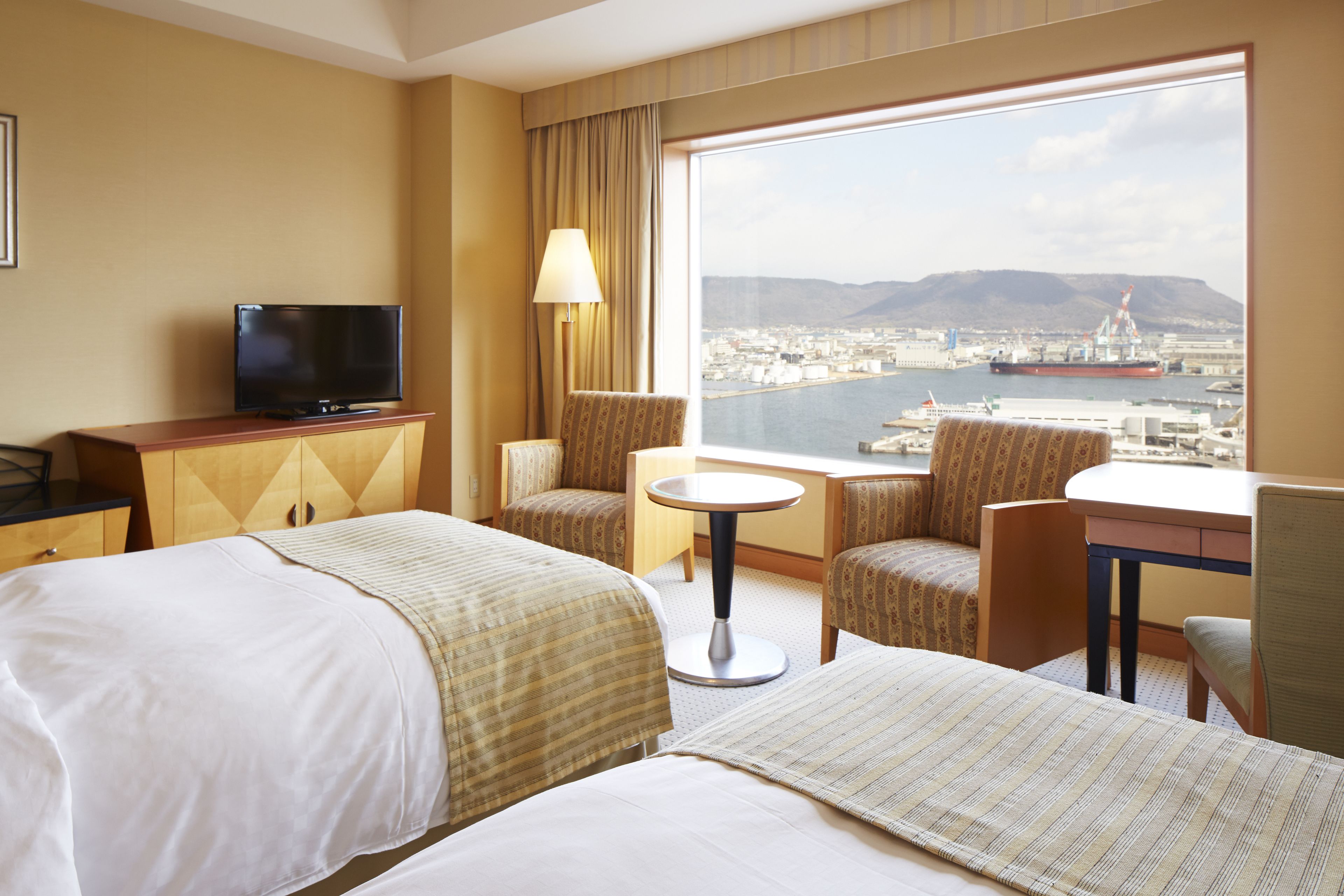 A hotel room with two beds, a television, a desk with a chair, a lamp, and a large window offering a view of a harbor and cityscape.