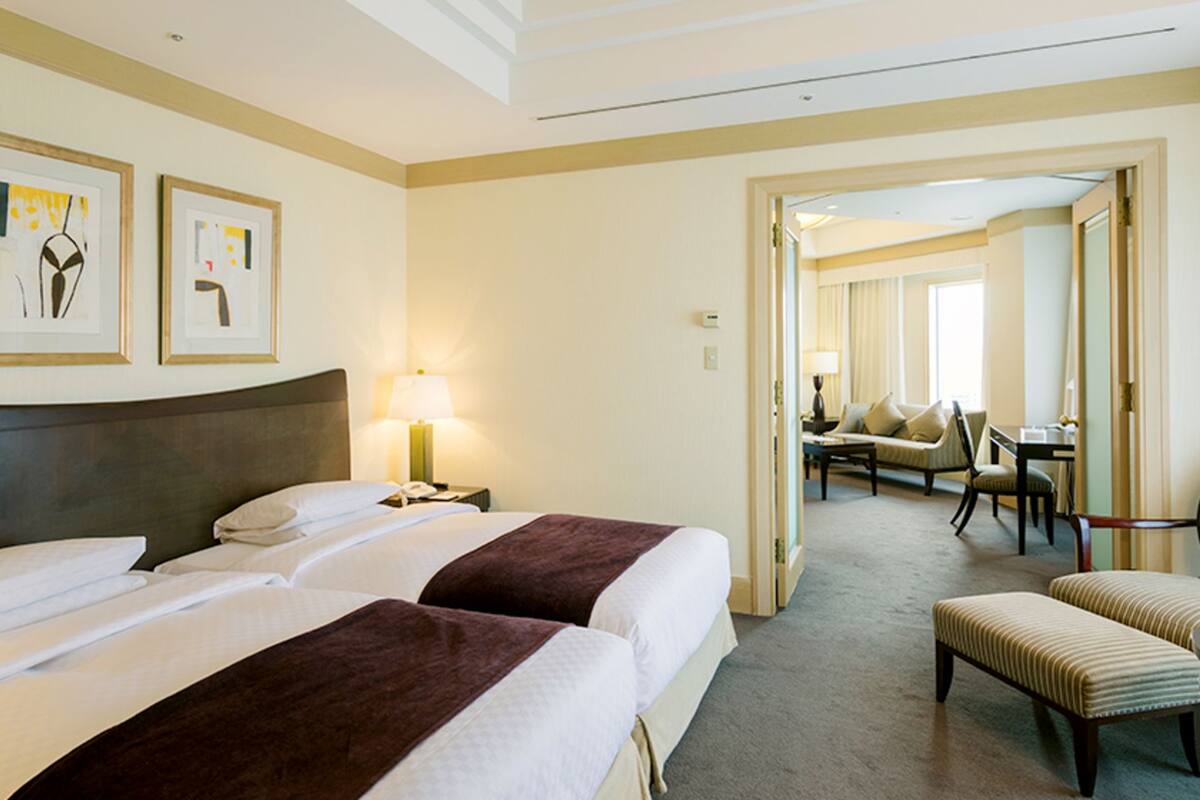 Habitación de hotel con dos camas, un área de estar con una silla y una mesa, y un pasillo que conduce a otra habitación.