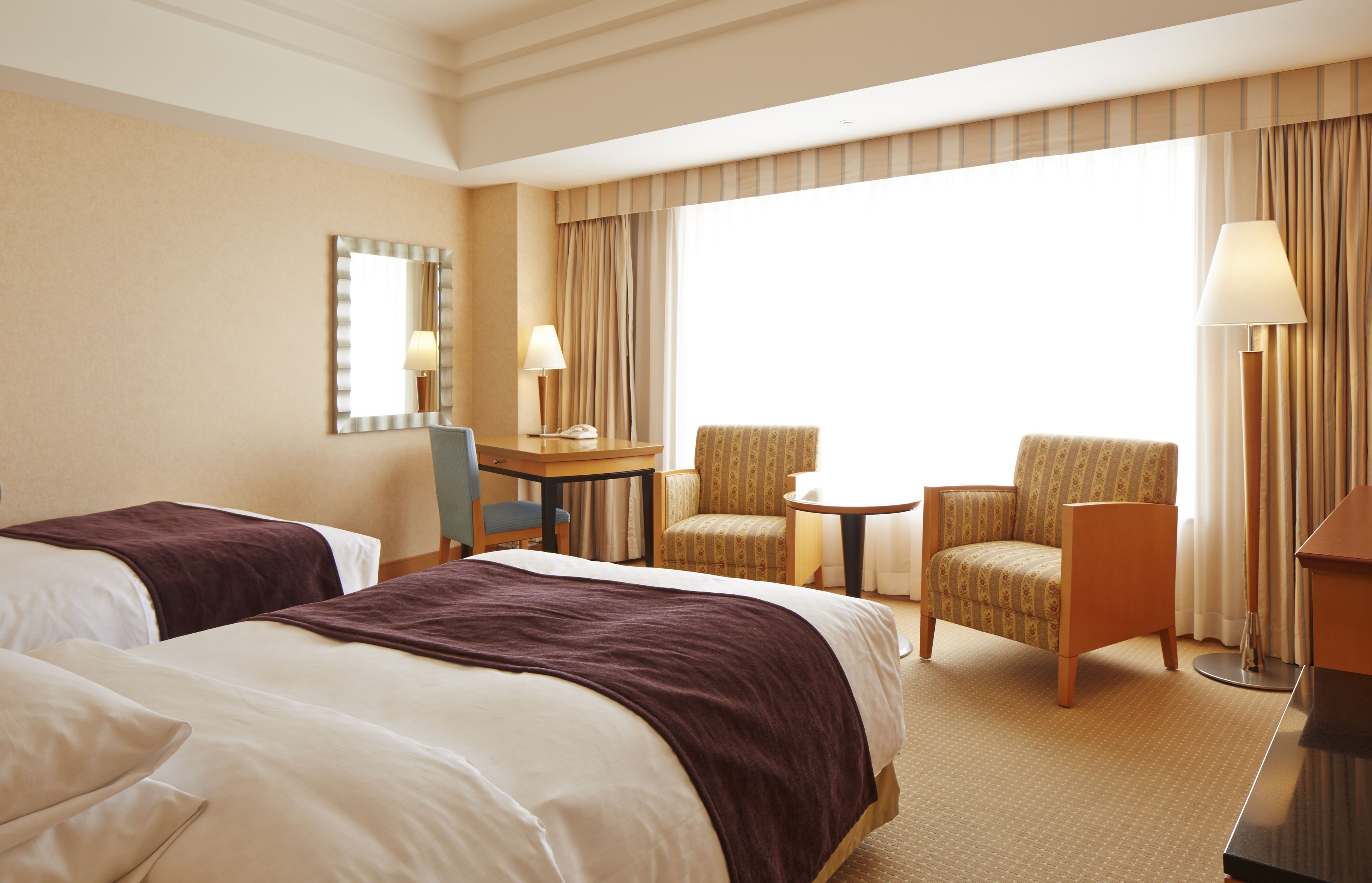A hotel room with two beds, a desk, two chairs, a lamp, and a large window with curtains.