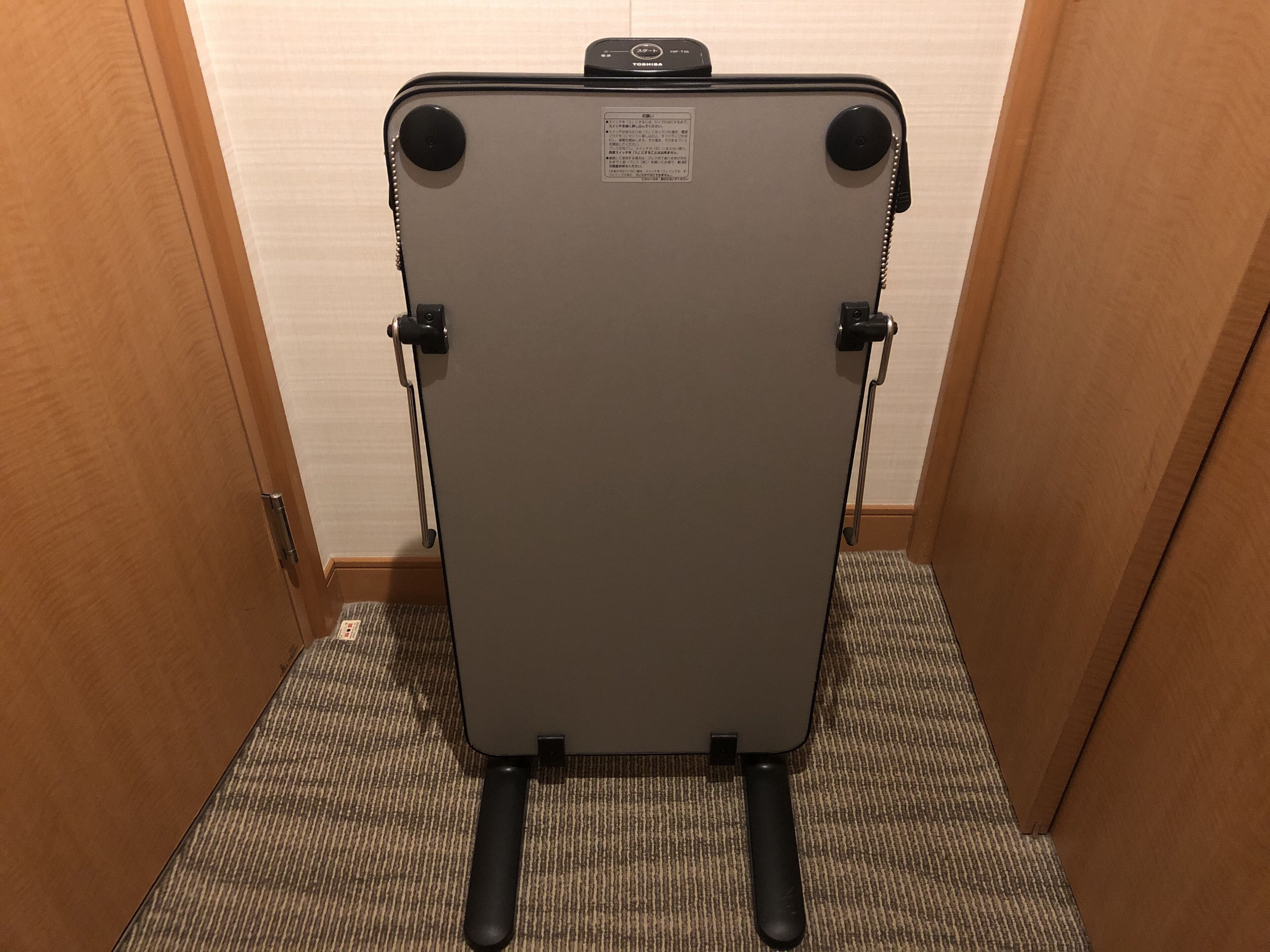 Una silla compacta gris, plegable, con asa y ruedas, ubicada en un pasillo estrecho con puertas de madera y suelo alfombrado.
