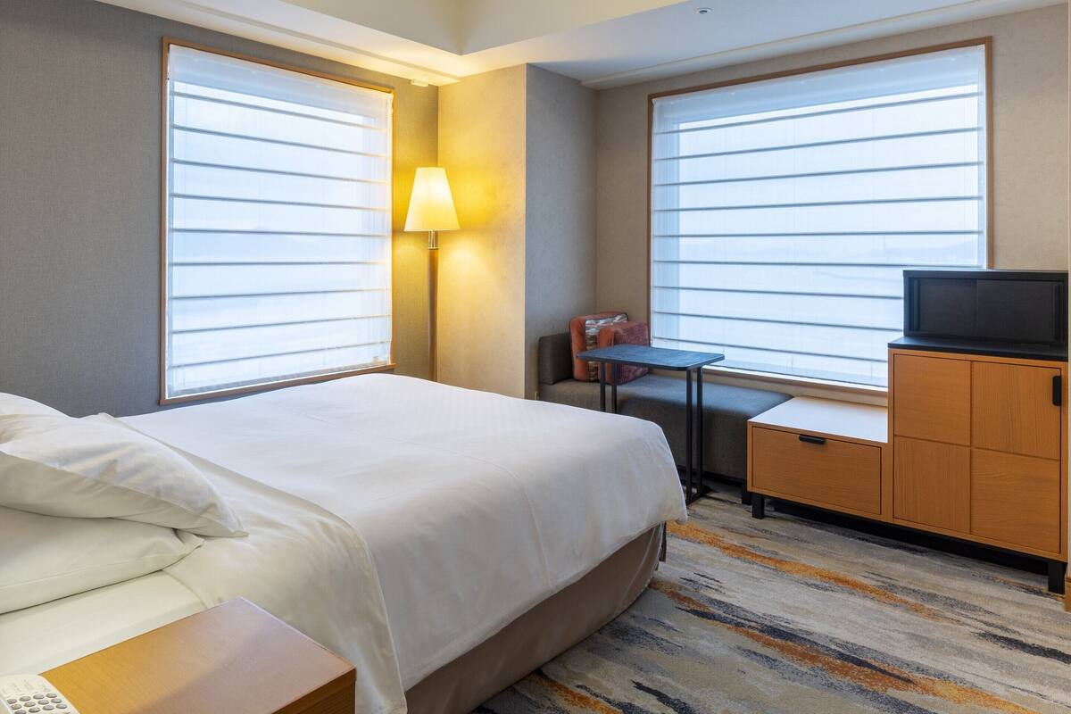 Habitación de hotel con cama, mesita de noche, lámpara, televisor y un pequeño escritorio con silla.