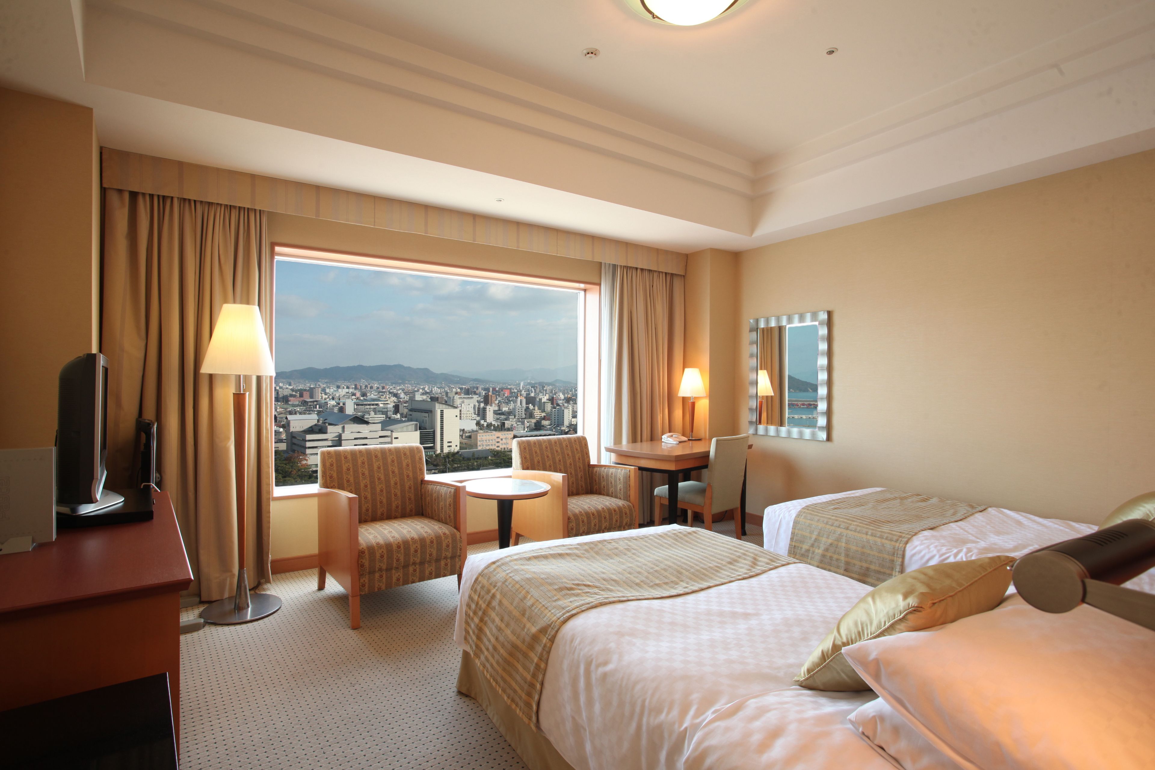 A hotel room with two beds, a desk, a chair, a small table, and a view of a cityscape.