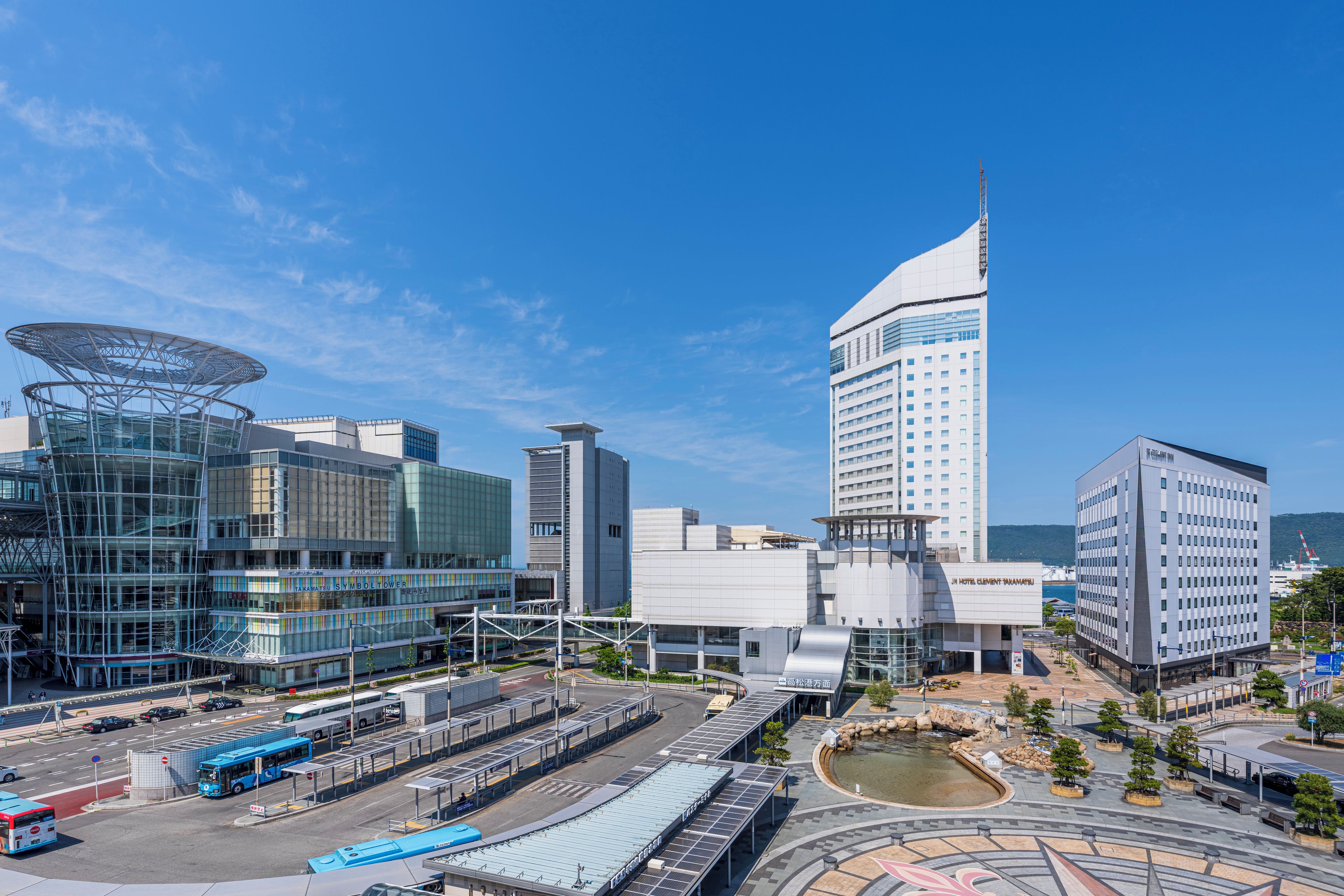 Uma área urbana moderna com uma torre imponente, um hotel e uma estação de ônibus.