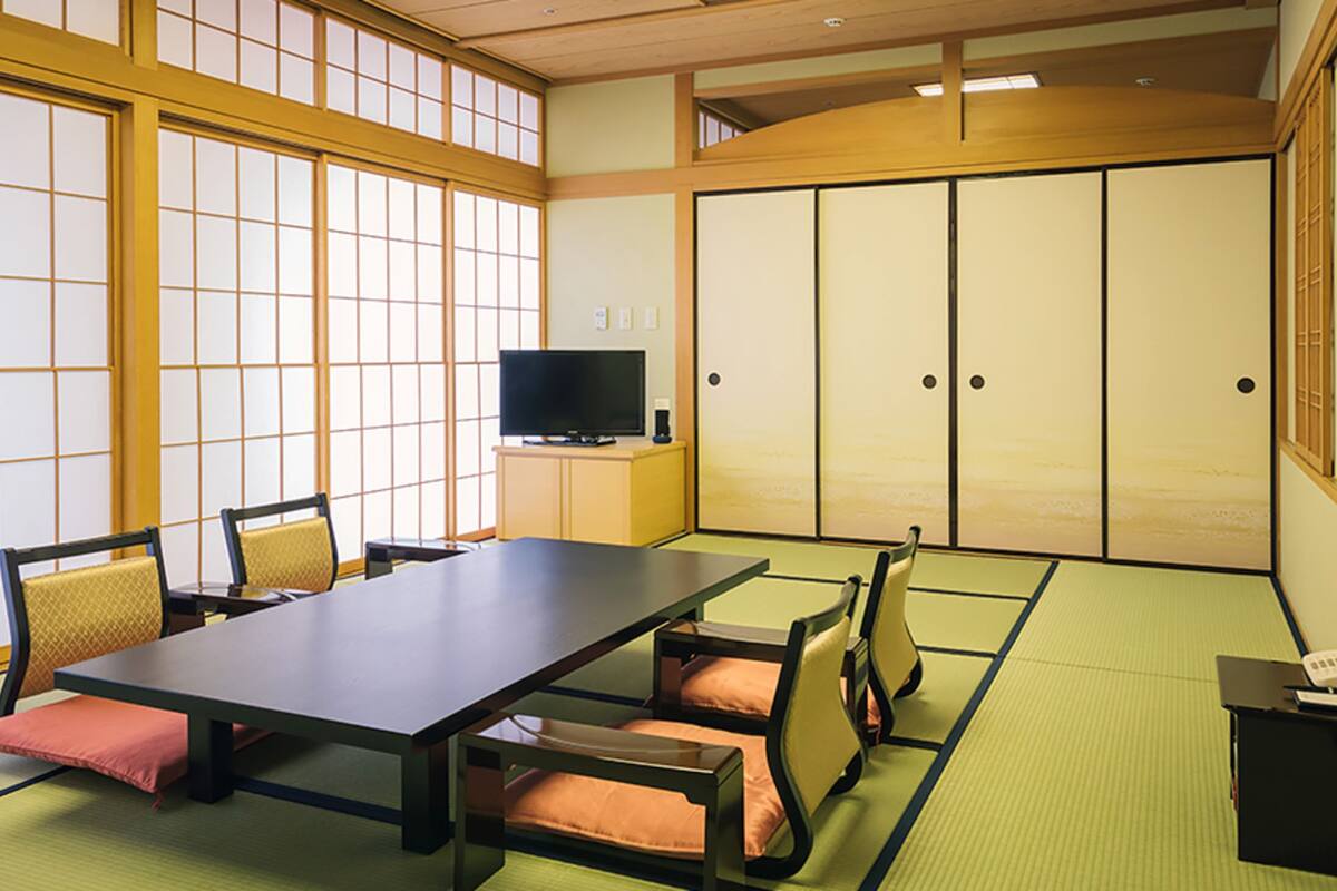 Una habitación tradicional japonesa con suelo de tatami, una mesa baja con cojines, un televisor sobre un mueble y puertas correderas.