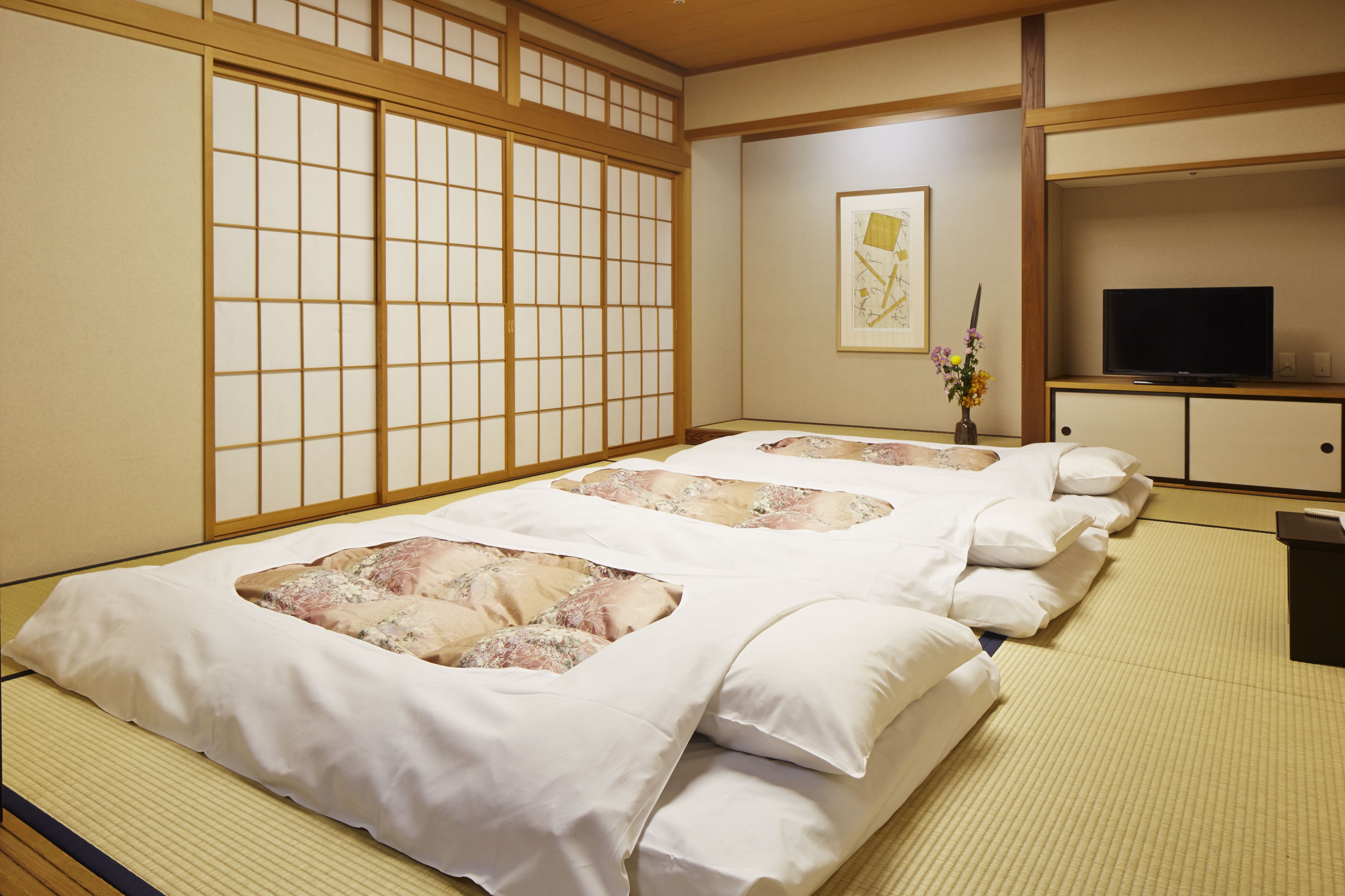 Una habitación japonesa tradicional con suelo de tatami, un futón, un televisor y un jarrón con flores.