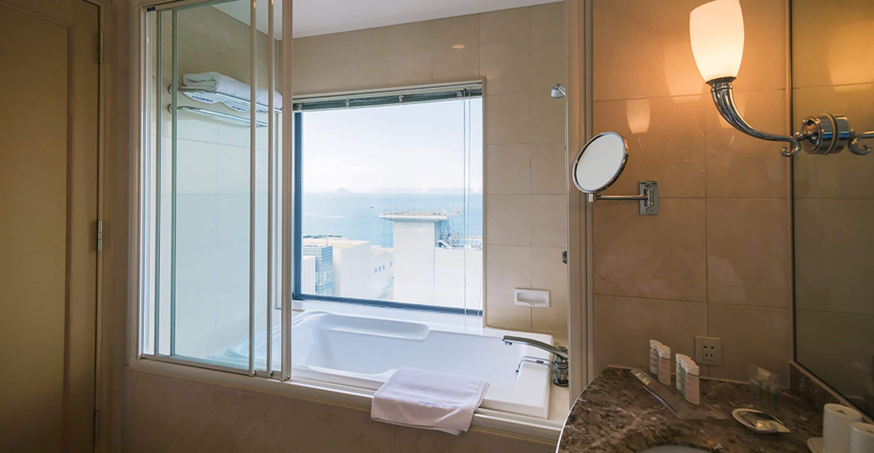 Une salle de bain moderne dotée d’une grande baignoire, d’une douche à parois de verre, d’un meuble-lavabo avec miroir et offrant une vue sur la ville par la fenêtre.