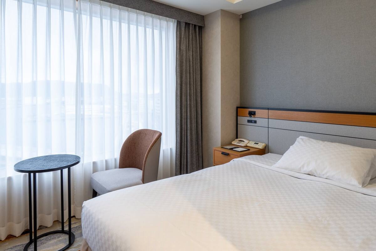 Habitación de hotel con cama, una silla, una mesita, un buró con teléfono y una ventana con cortinas.