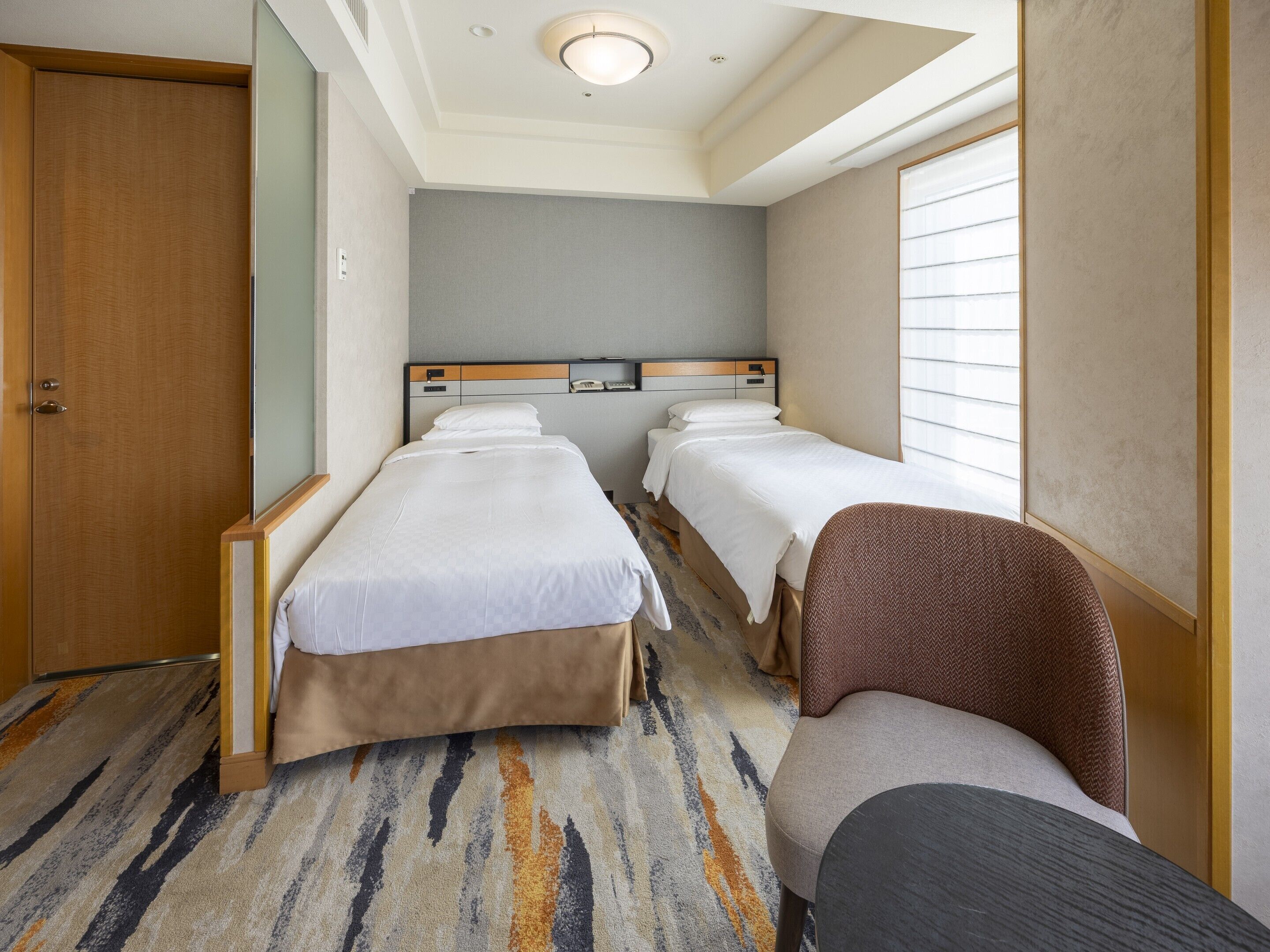 A hotel room with two beds, a wooden wardrobe, a carpeted floor, and a window with blinds.