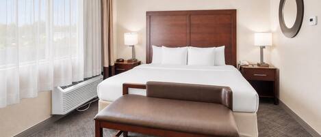 Una habitación de hotel con una cama grande, dos mesitas de noche con lámparas, un banco, un ventanal con cortinas y un espejo colgado en la pared.
