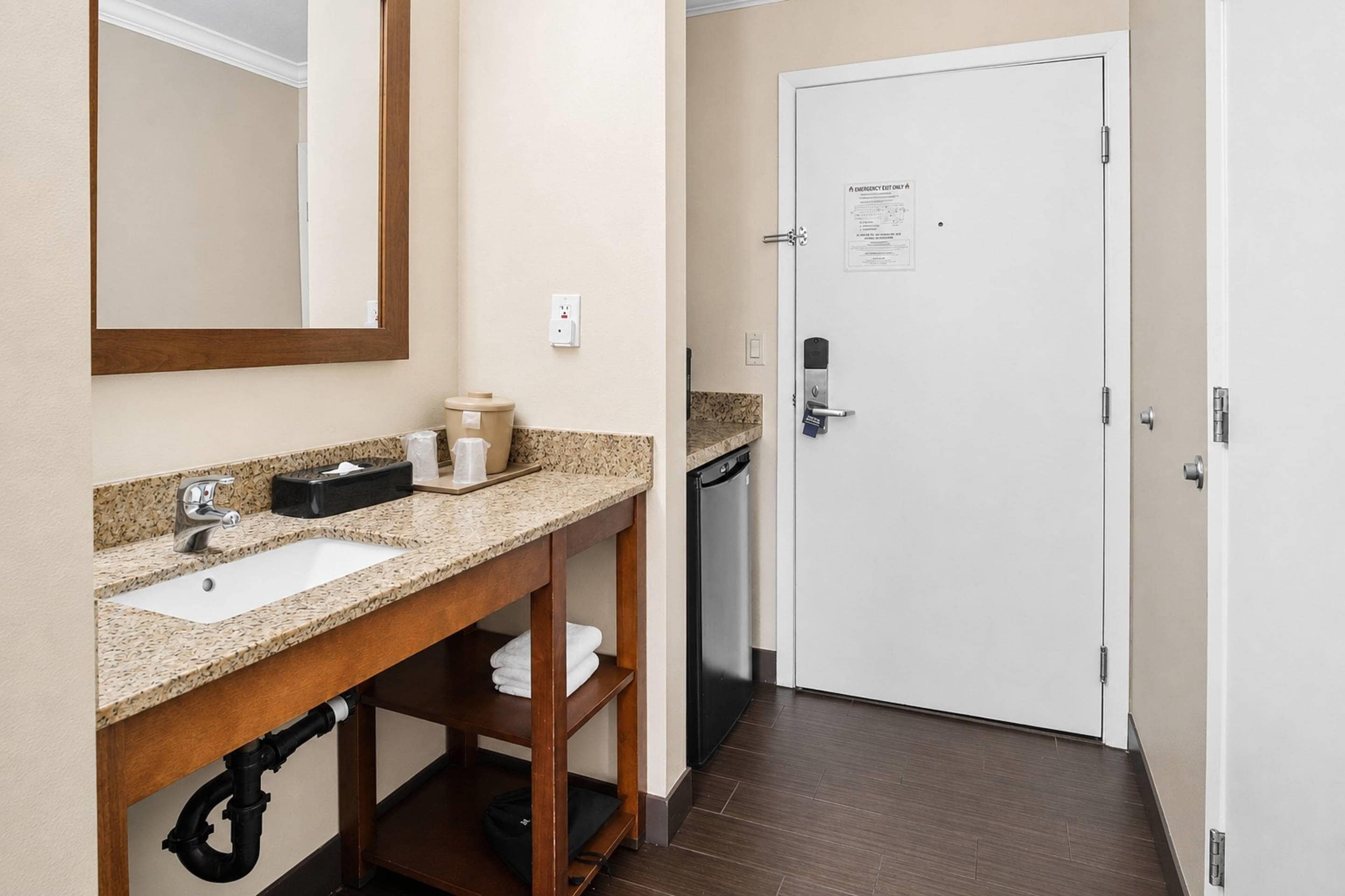 Un baño de hotel con lavamanos, espejo y una puerta con letrero de salida de emergencia.