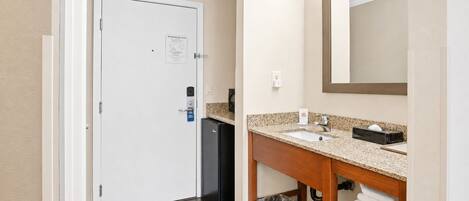 A hotel bathroom with a white door, a sink with a granite countertop, a mirror, and a trash bin.