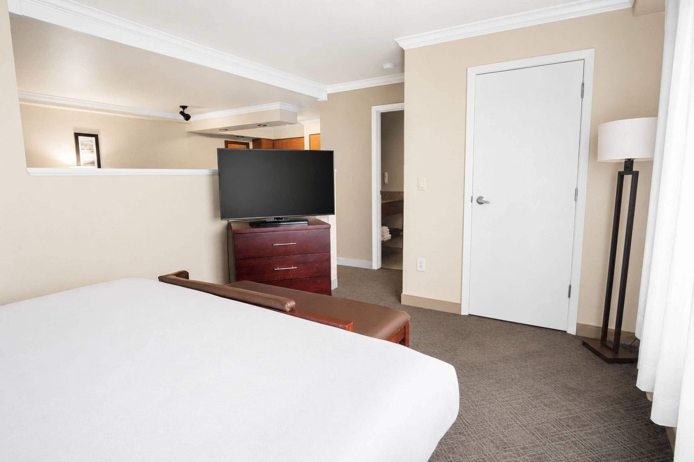 Ein Hotelzimmer mit einem großen Bett, einem Fernseher auf einer Holzkommode, einer Tür und einer Stehlampe.