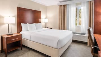 A hotel room with a large bed, two bedside lamps, a desk with a chair, a window with curtains, and a wall-mounted heater.