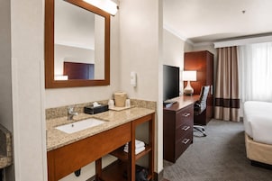 Habitación de hotel con tocador, cama, escritorio con silla, televisión y ventana con cortinas.
