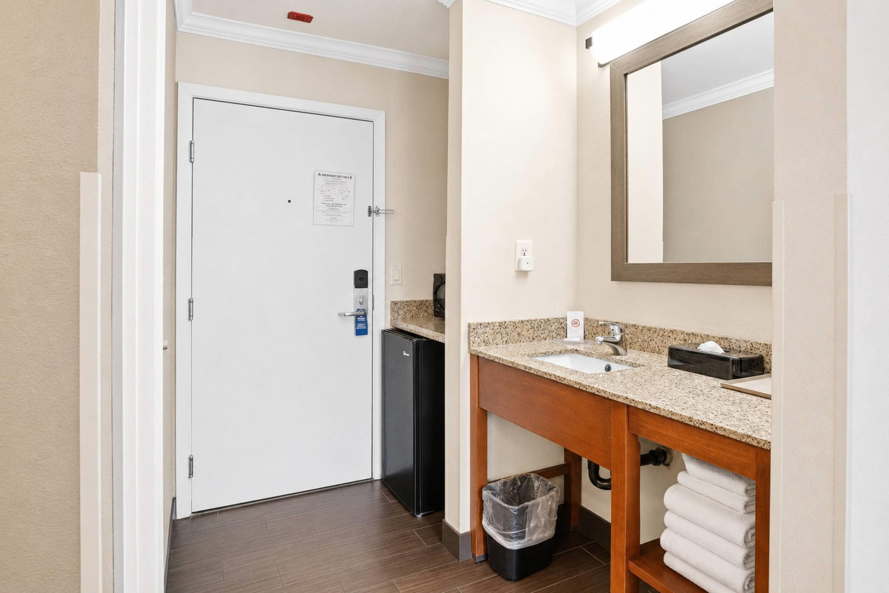 Un baño de hotel con una puerta blanca, un lavabo con espejo, un cesto de basura y un perchero para toallas.
