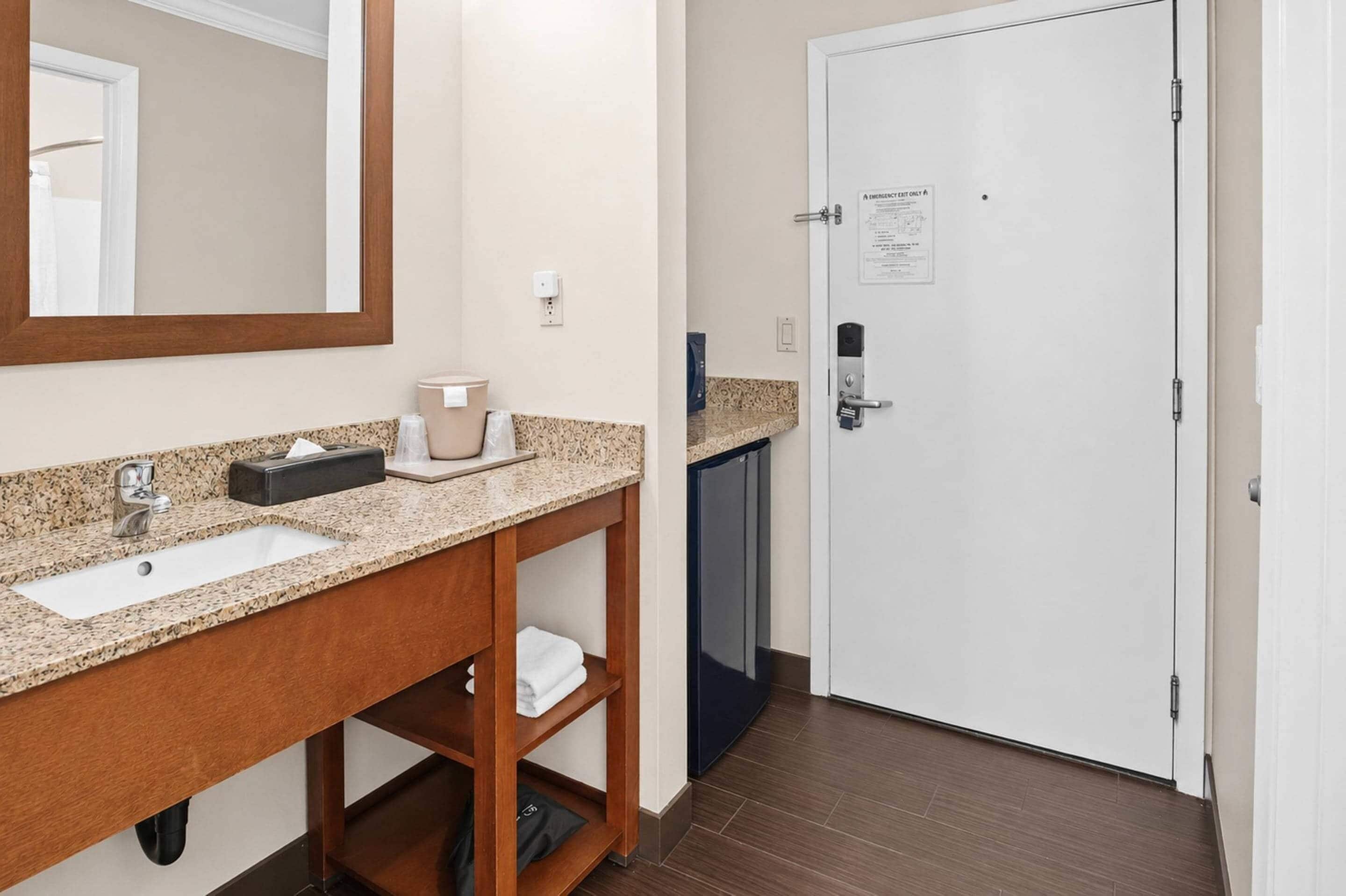 A hotel bathroom with a sink, mirror, towel rack, and a door with an emergency exit sign.